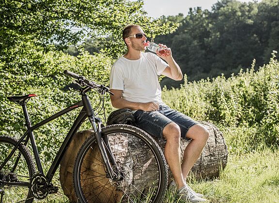 Radfahrer mit Sonnenbrille trinkt Wasser aus einer Flasche während einer Pause auf einem Baumstamm. Sein schwarzes Mountainbike steht daneben.